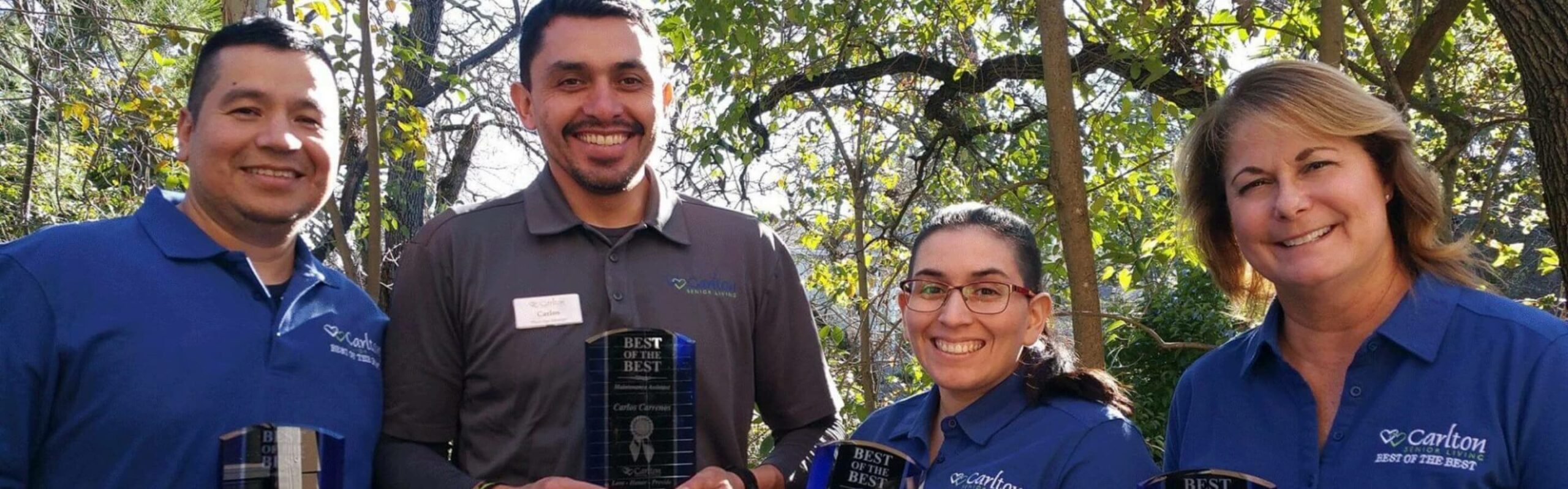 Four Carlton Senior Living team members, smiling proudly outdoors in blue uniforms, showcasing an award titled 'Best of the Best.' Greenery in the background highlights the beautiful environment of the community.