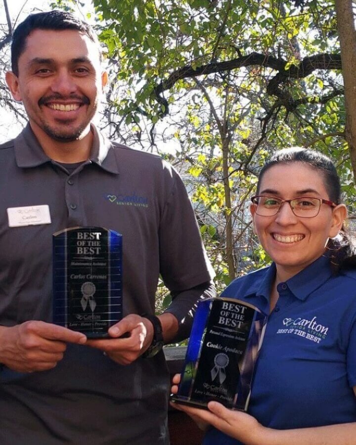 Four Carlton Senior Living team members, smiling proudly outdoors in blue uniforms, showcasing an award titled 'Best of the Best.' Greenery in the background highlights the beautiful environment of the community.