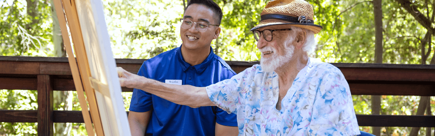 An elderly man with a straw hat and a floral shirt paints on a canvas, guided by a smiling Carlton team member in a blue uniform, exemplifying the engaging activities provided at Carlton Senior Living.