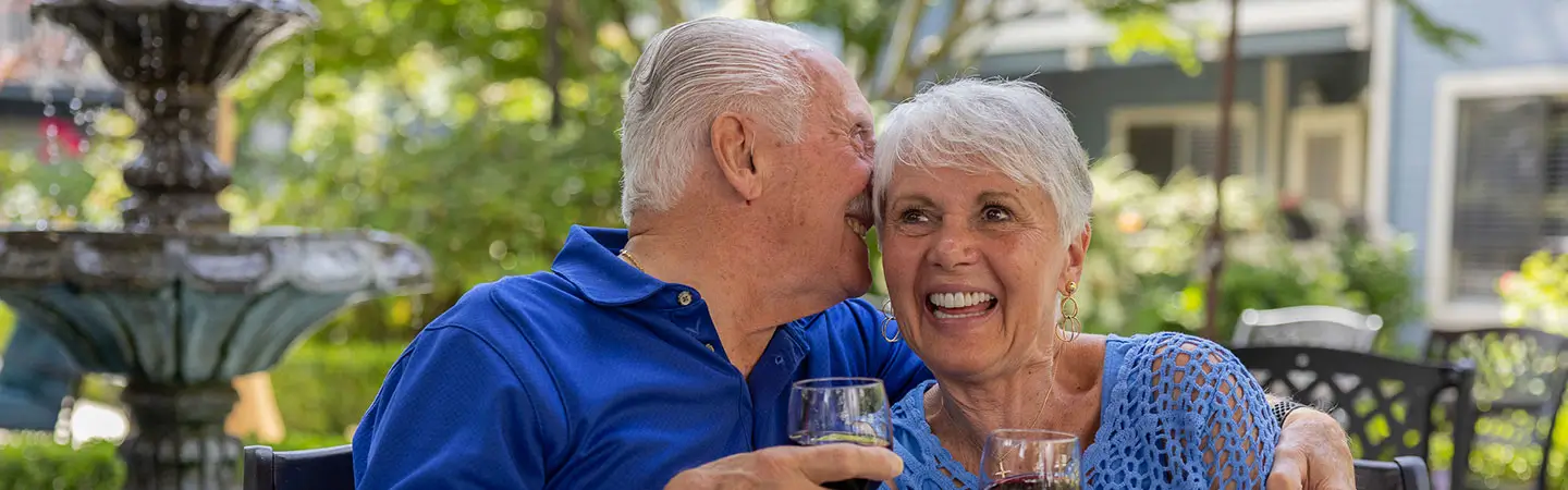 An elderly couple hold wine glasses and sit by a fountain, sharing a happy moment at Carlton Senior Living.