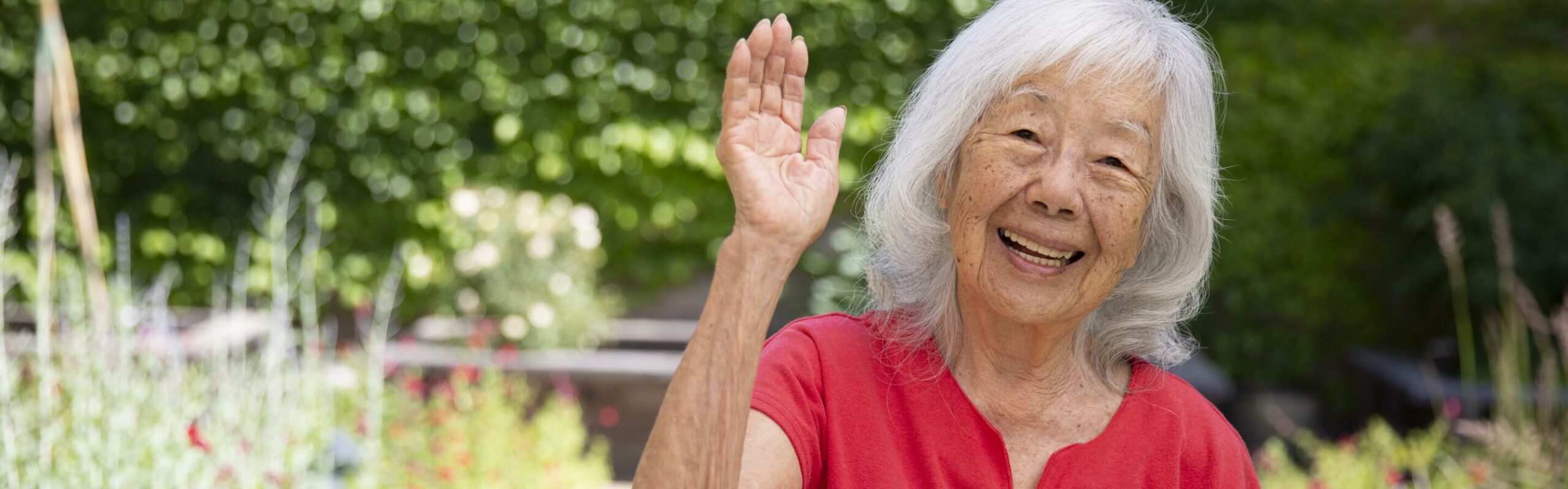 An elderly woman with silver hair and a radiant smile waves cheerfully in a garden, conveying the friendly community atmosphere at Carlton Senior Living.