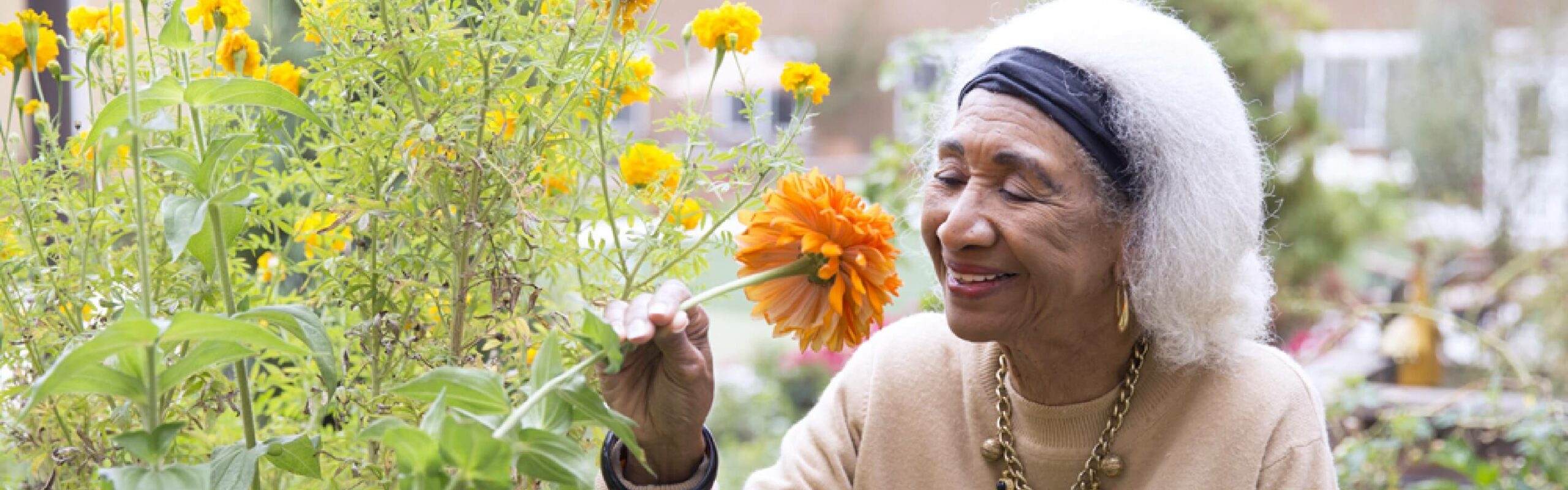 An elderly woman with white hair and a headband smells an orange flower in a lush garden, wearing a beige sweater and gold jewelry, illustrating the serene outdoor experiences available at Carlton Senior Living.