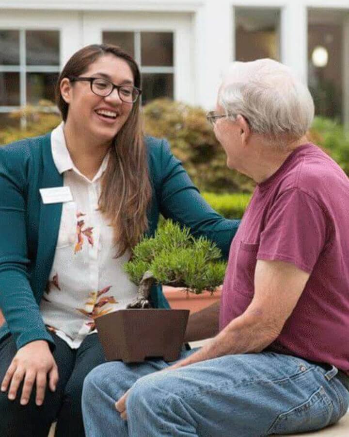 An elderly man and a Carlton team member share a laugh while seated on a bench by a fountain, representing the caring and friendly team interactions at Carlton Senior Living.