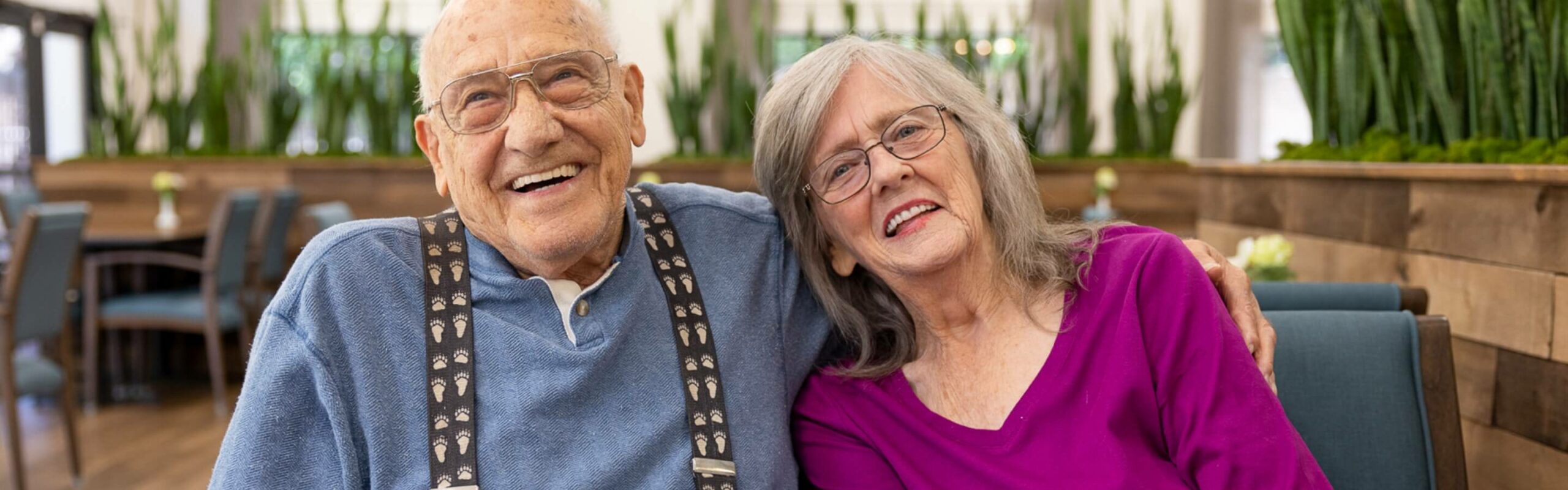 An elderly couple sitting together; the man is laughing and the woman leans on his shoulder smiling, demonstrating the community atmosphere at Carlton Senior Living.