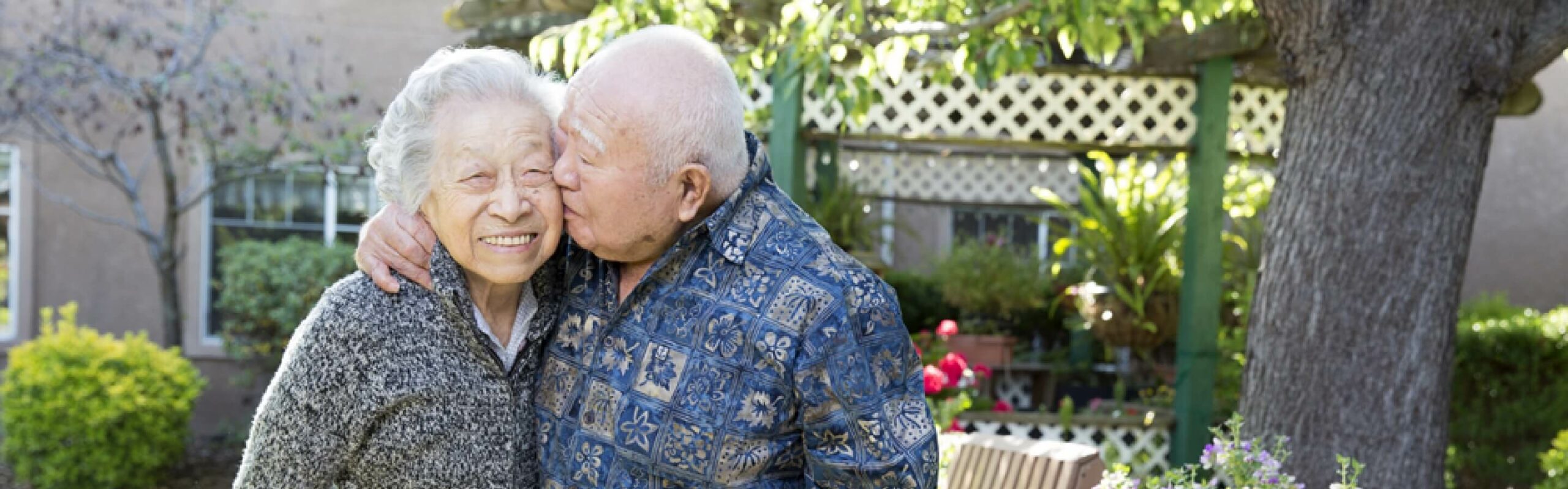 An elderly couple, with the man giving a kiss on the cheek to a woman, both smiling in a garden at Carlton Senior Living.