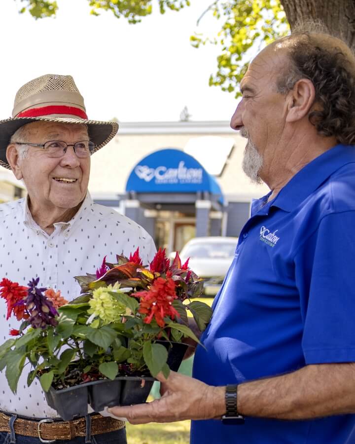 An elderly man in a hat and white shirt smiling by colorful plants accompanied by a Carlton team member in a blue shirt, showcasing the community's outdoor setting.