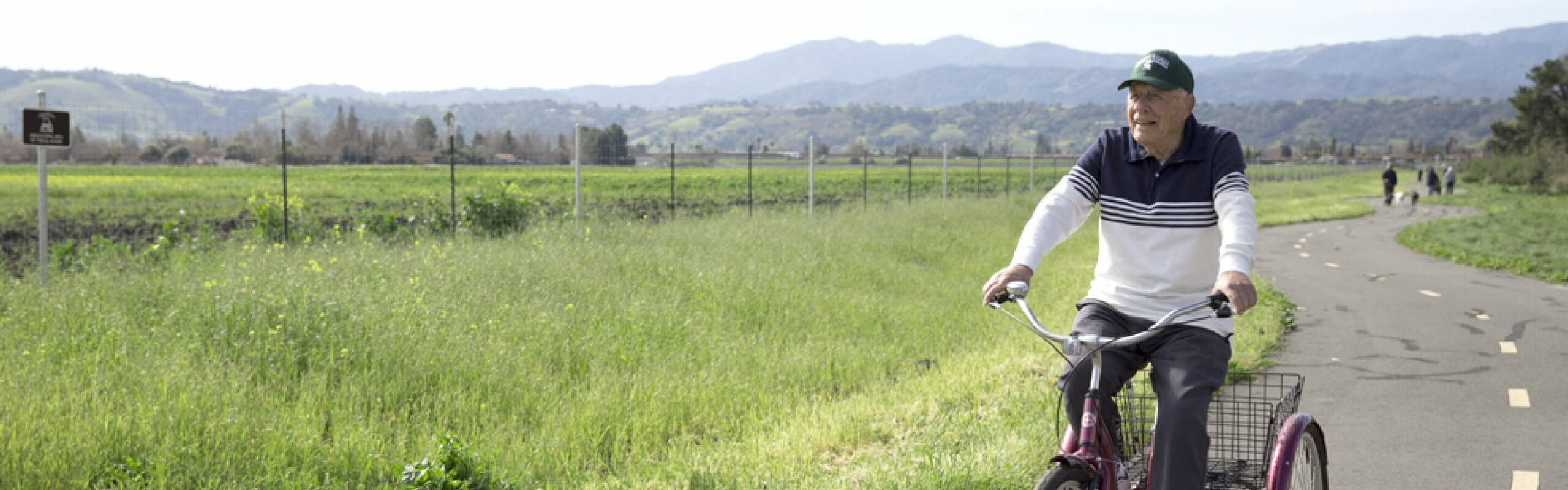 An elderly man enjoys a bicycle ride along a winding road, admiring green fields and mountain views, depicting an active lifestyle at Carlton Senior Living.