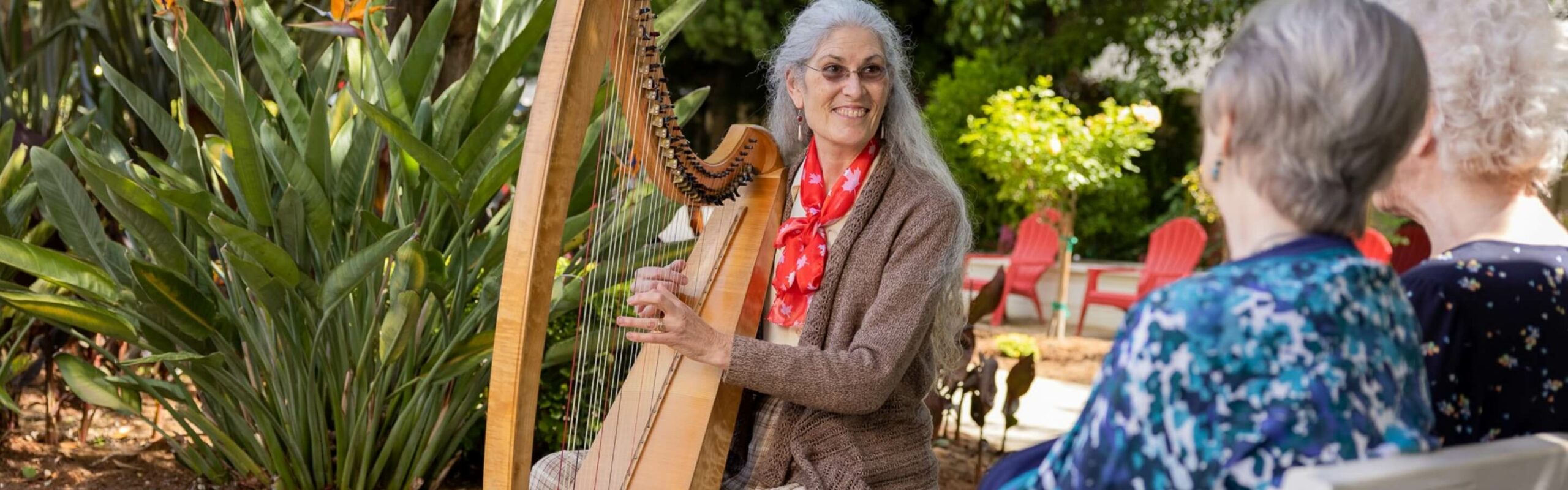 An elderly woman with long gray hair, playing a harp in a garden for an audience, creates a serene musical setting at Carlton Senior Living.