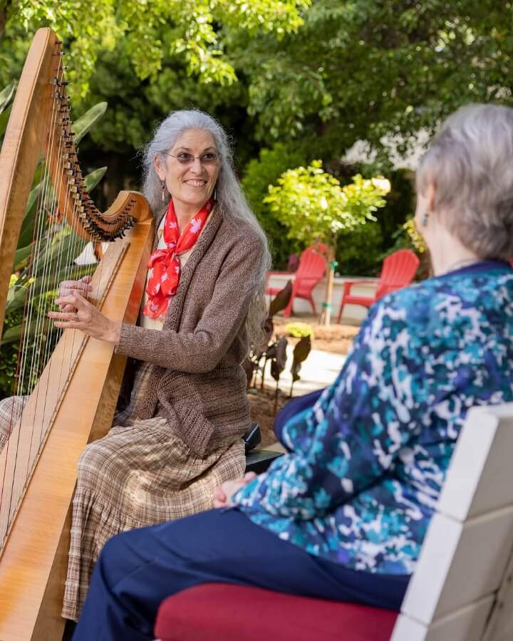 An elderly woman with long gray hair, playing a harp in a garden for an audience, creates a serene musical setting at Carlton Senior Living.