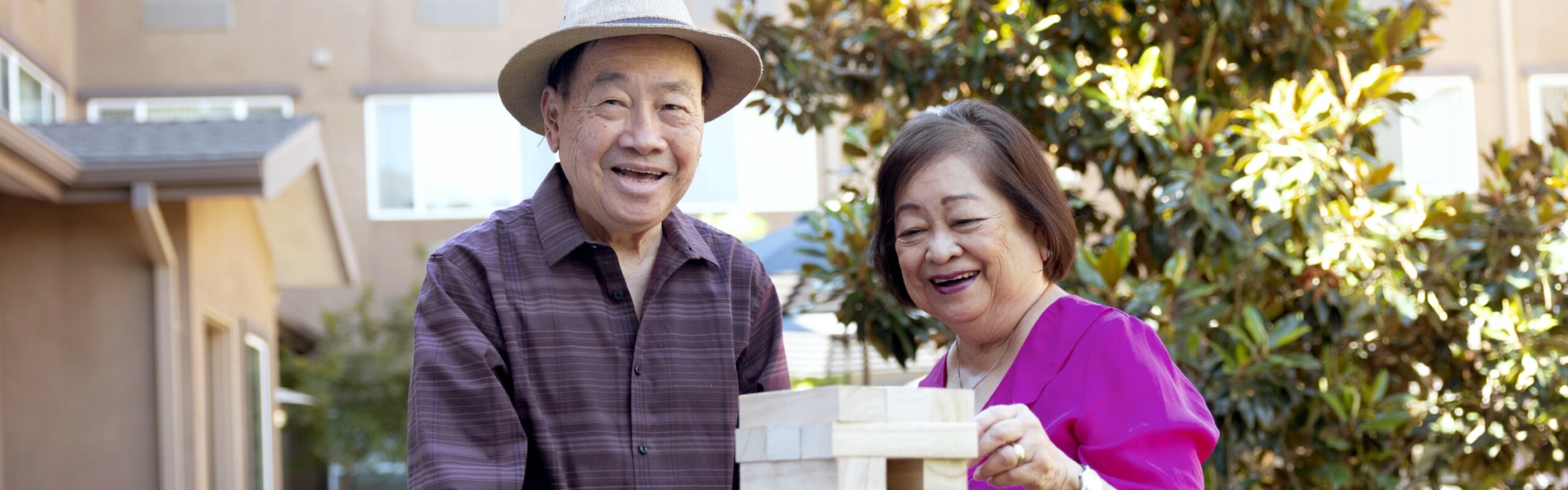 An elderly couple laugh together while playing a game outdoors, with the man in a hat and striped shirt and the woman in a fuchsia top, embodying the active and social lifestyle at Carlton Senior Living.