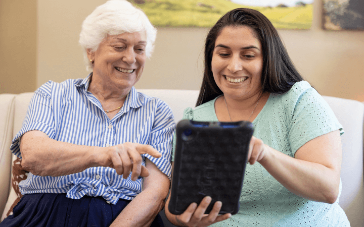 An elderly woman and a Carlton Senior Living staff member smiling while using a tablet together, highlighting innovative technology in senior care.